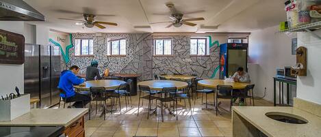 A kitchen with two ceiling fans, a refrigerator, and a microwave. There are two dining tables with chairs.