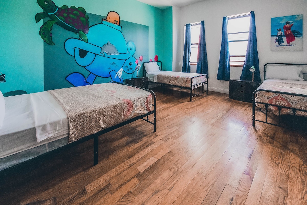 A room with three beds, wooden flooring, a mural on the wall, and a window with curtains.