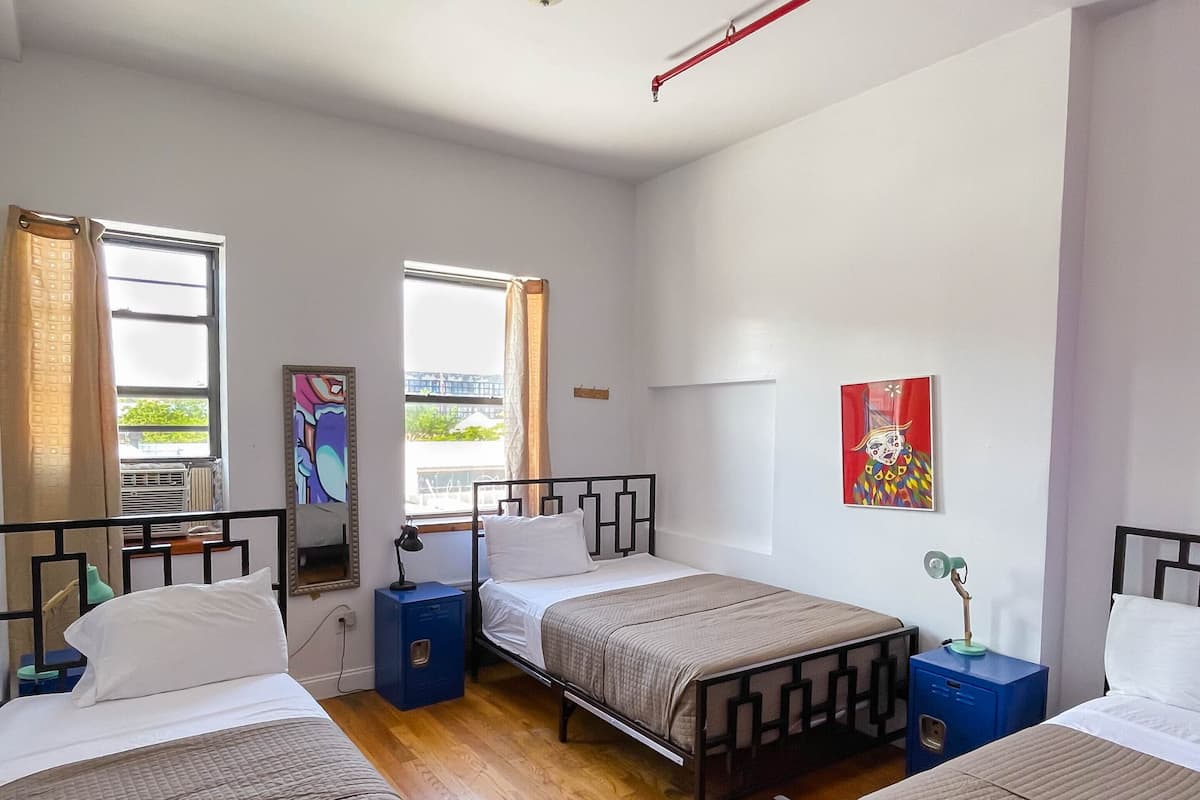 A room with three beds, a window with curtains, a painting on the wall, and a ceiling light fixture.