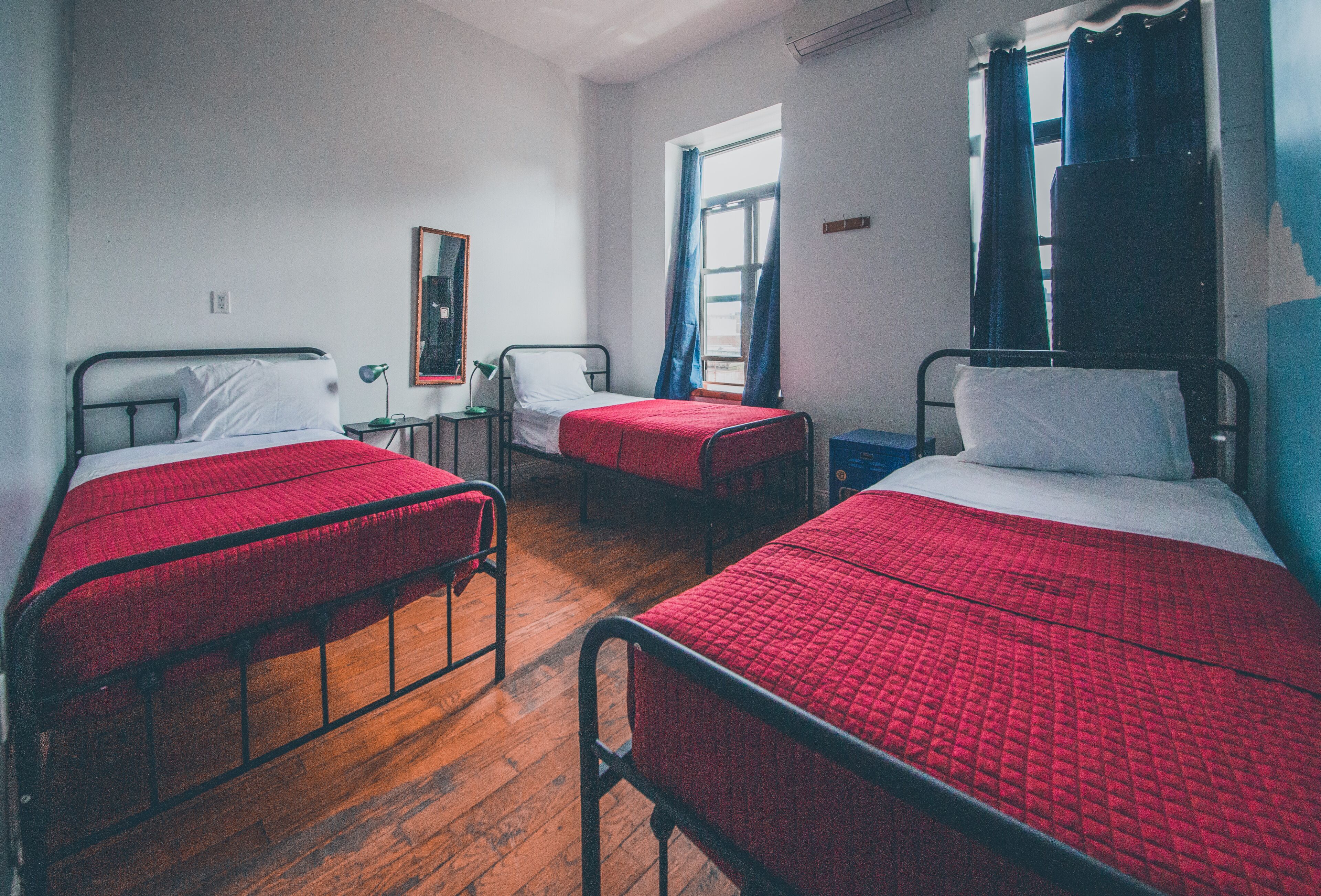A dormitory room with three single beds, each with a red quilted bedspread and white pillows. There is a small table with a lamp, a window with blue curtains, and a mirror on the wall.