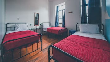 A dormitory room with three single beds, each with a red quilted bedspread and white pillows. There is a small table with a lamp, a window with blue curtains, and a mirror on the wall.
