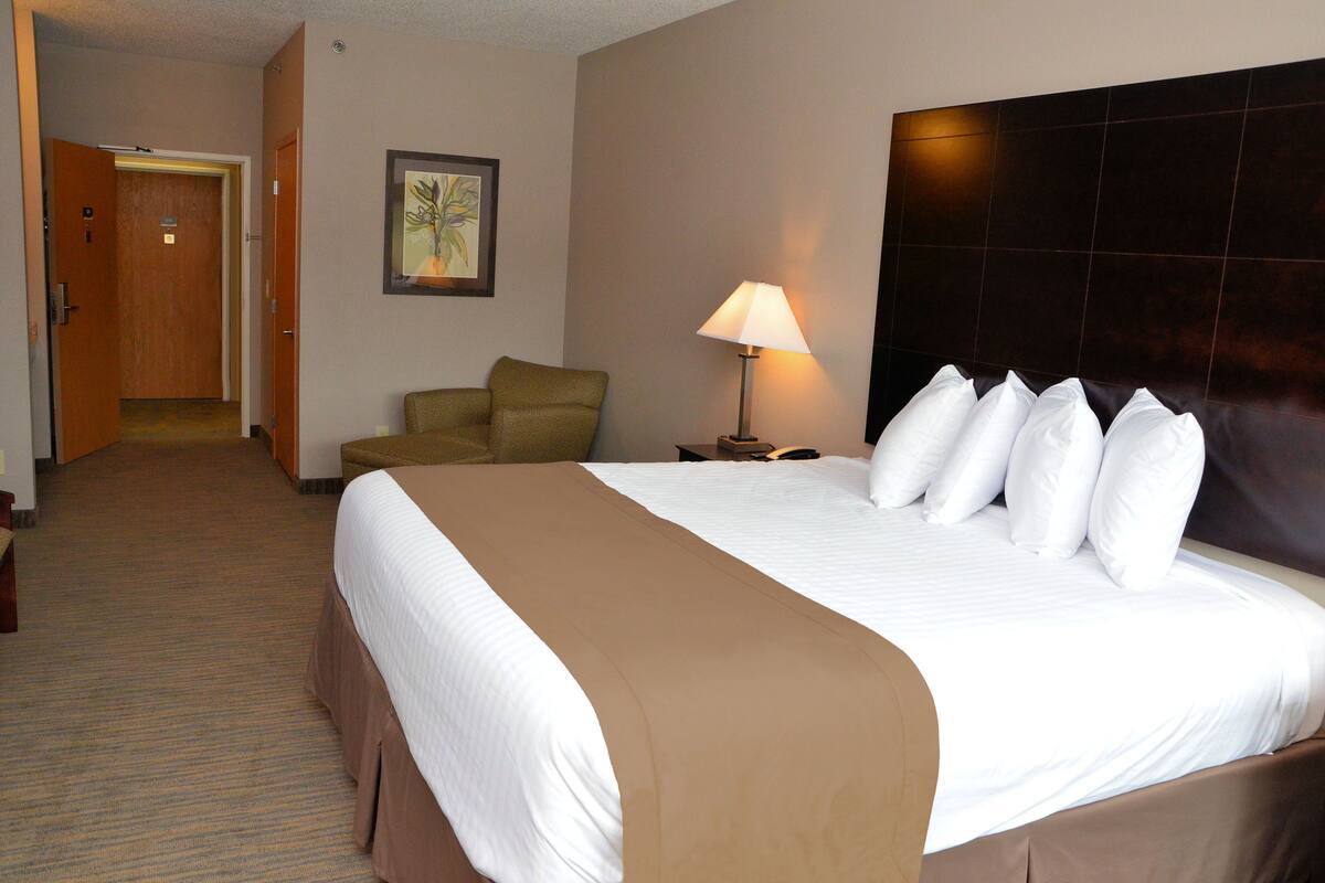 Habitación de hotel con una cama grande, una silla, una mesita de noche con lámpara y una puerta que da a otra habitación.