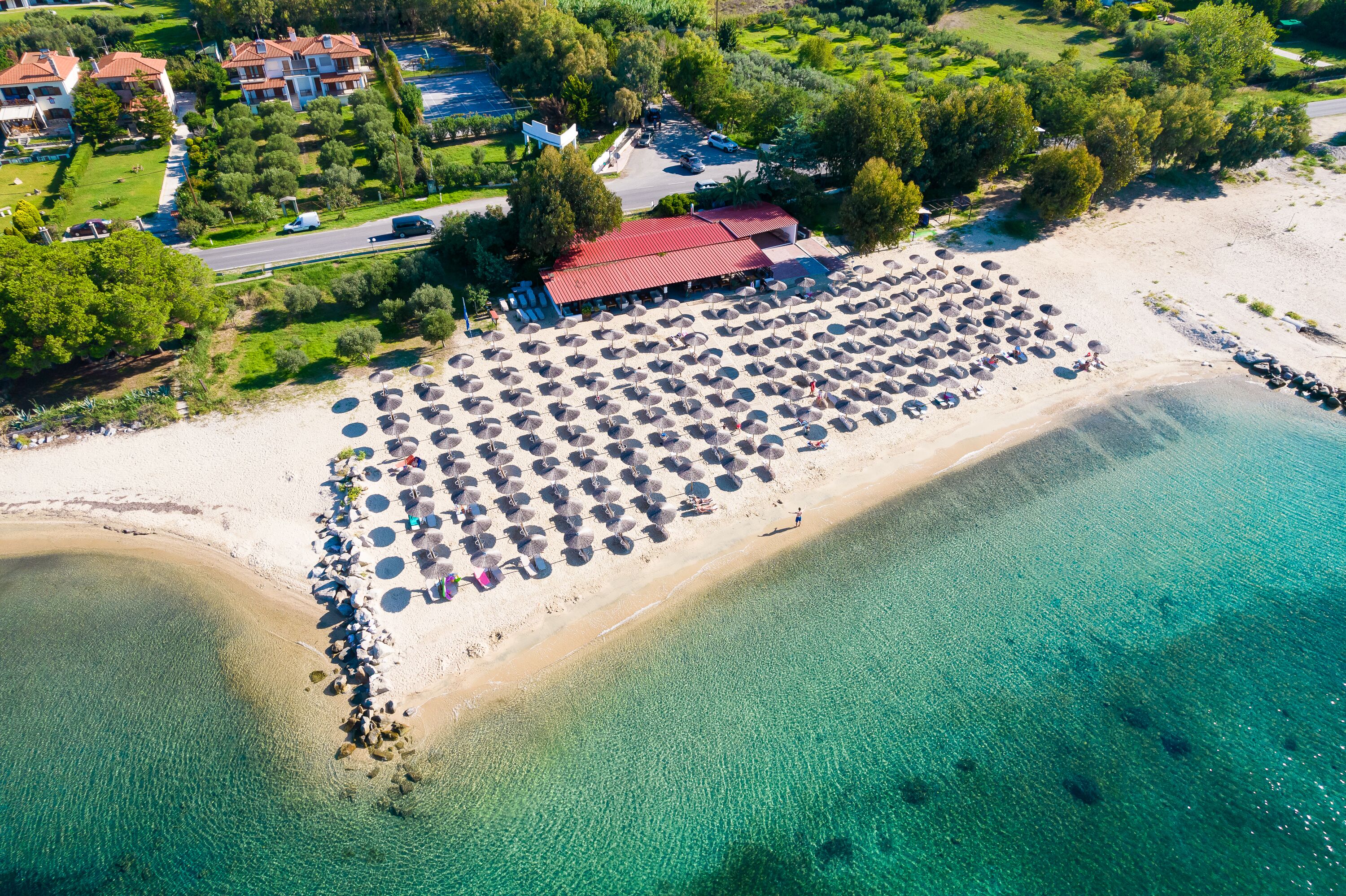 An aerial view of a beach resort with a large number of umbrellas and lounge chairs, a building with a red roof, and a clear view of the turquoise sea.