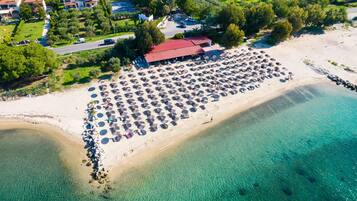 An aerial view of a beach resort with a large number of umbrellas and lounge chairs, a building with a red roof, and a clear view of the turquoise sea.