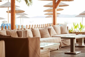 A beachside lounge with striped sofas, a glass-top table, and thatched umbrellas.