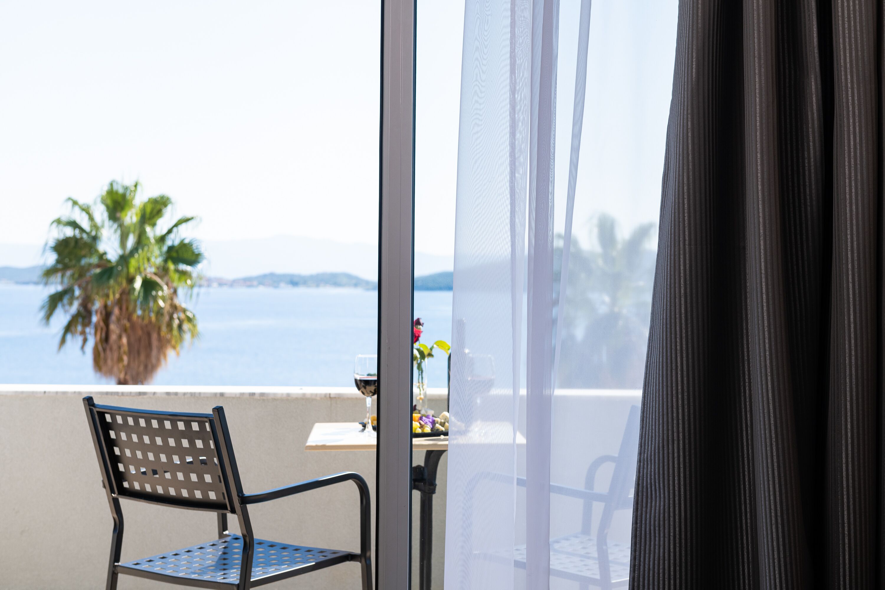 A balcony with a metal chair, a small table with a wine glass and flowers, and a view of the sea and palm trees.