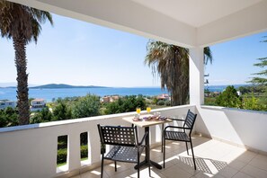 Un balcón con mesa y sillas, con vistas a la costa, casas y palmeras.