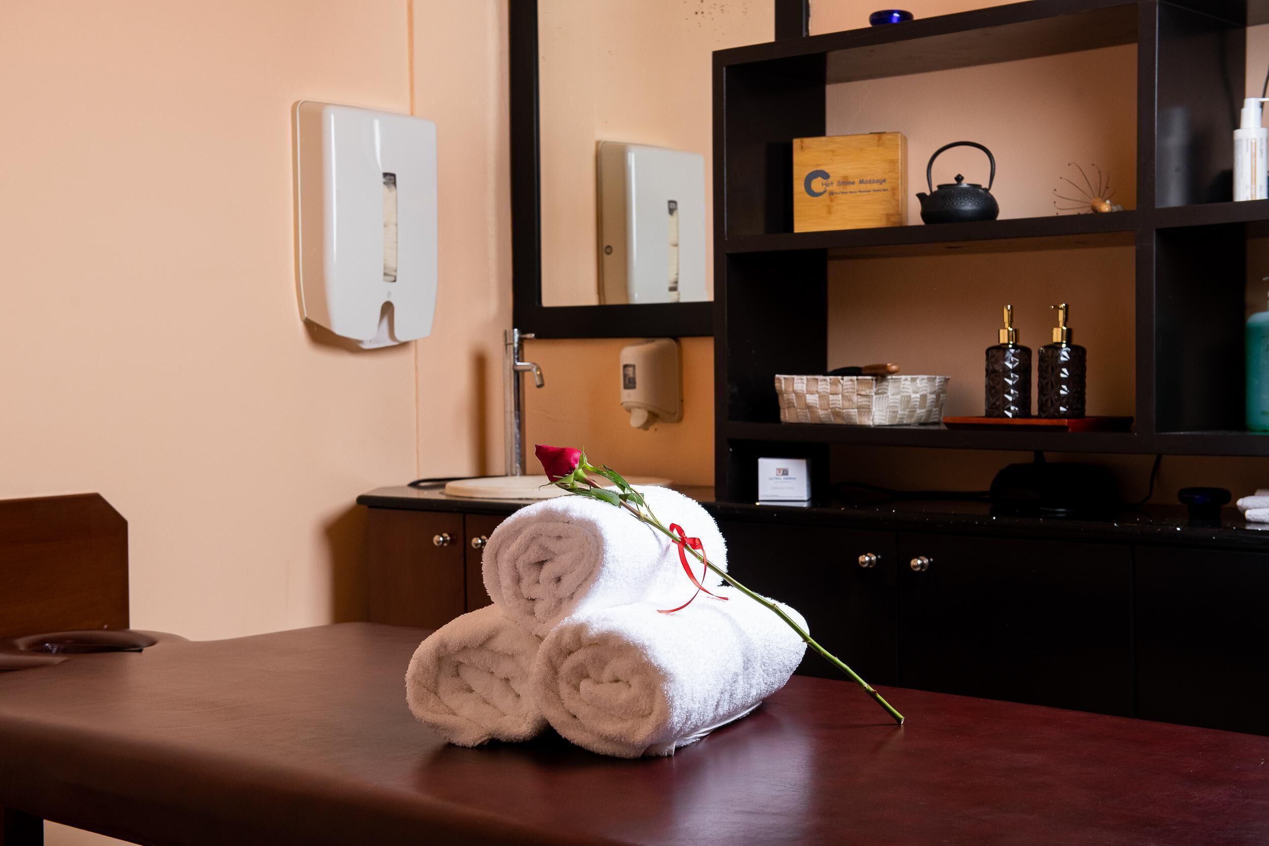 Hotelkamer met een houten bankje, een opgerolde handdoek, een roos, een spiegel, een plank met toiletartikelen en een doosje met de tekst "Cocacola".