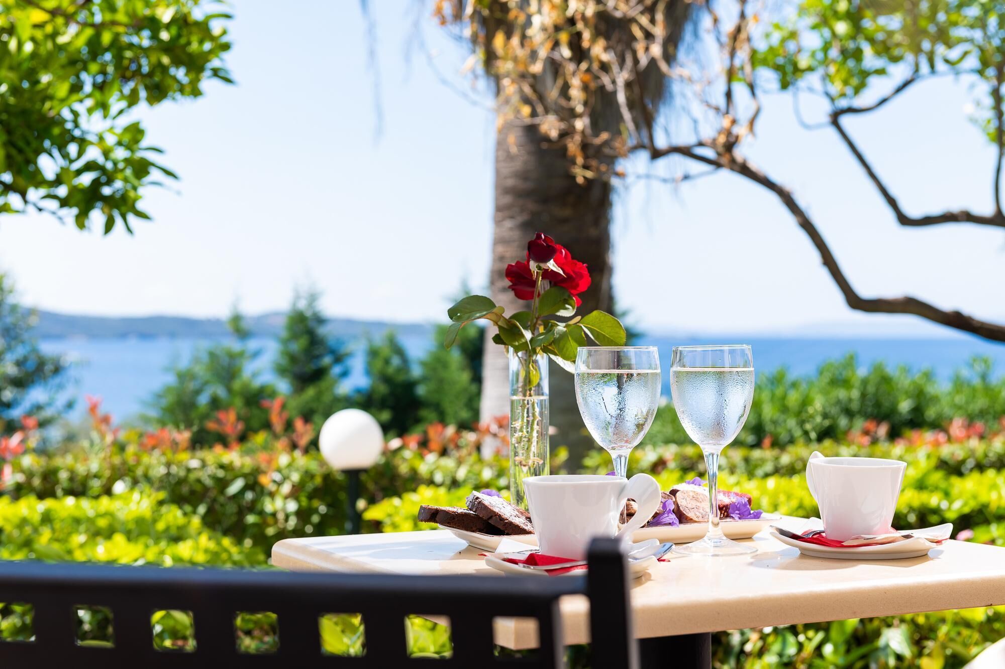 Ein Tisch im Freien mit Blick auf das Meer, eine Vase mit roten Blumen und zwei Gläser Wasser.