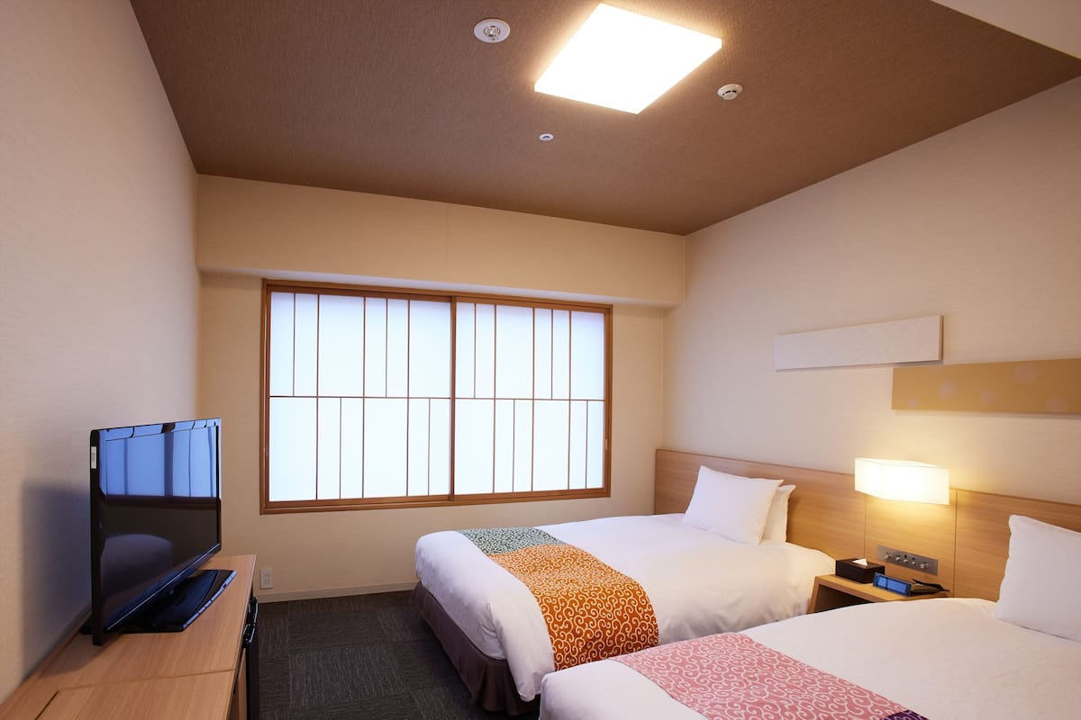 A hotel room with two beds, a TV, a desk, and a window with blinds.