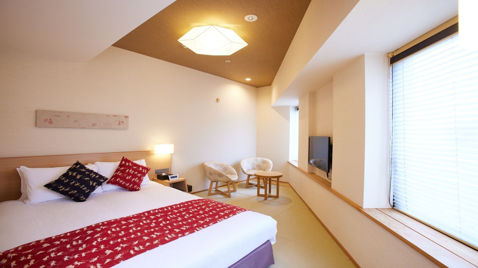 A hotel room with a bed, two chairs, a TV, and a window with blinds.