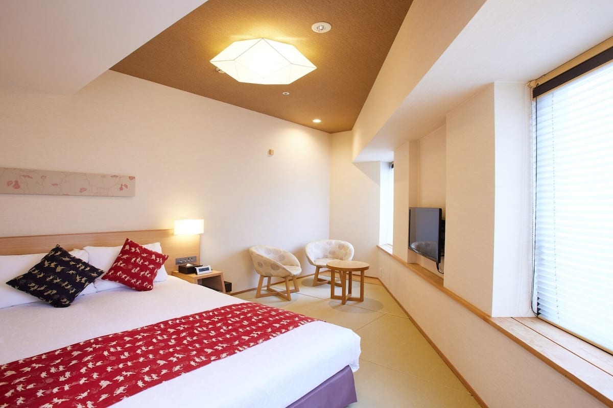 A hotel room with a bed, two chairs, a TV, and a window with blinds.