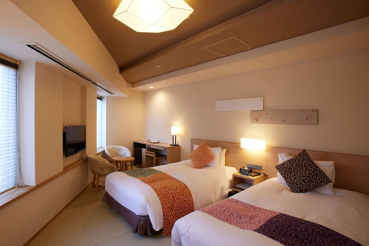 A hotel room with two beds, a desk, a chair, and a TV.