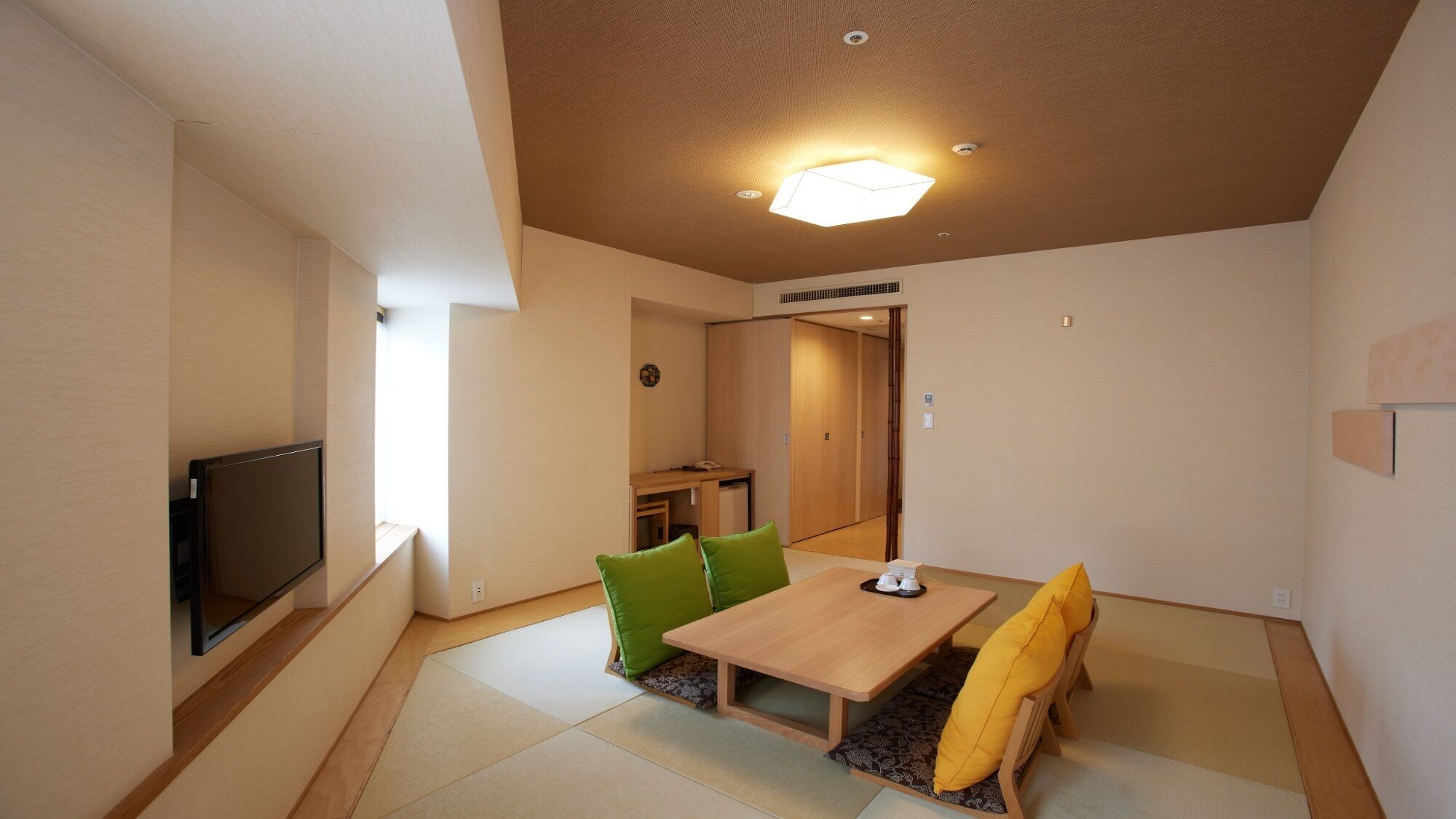 A room with a TV, a wooden table, green and yellow cushions, and a ceiling light.