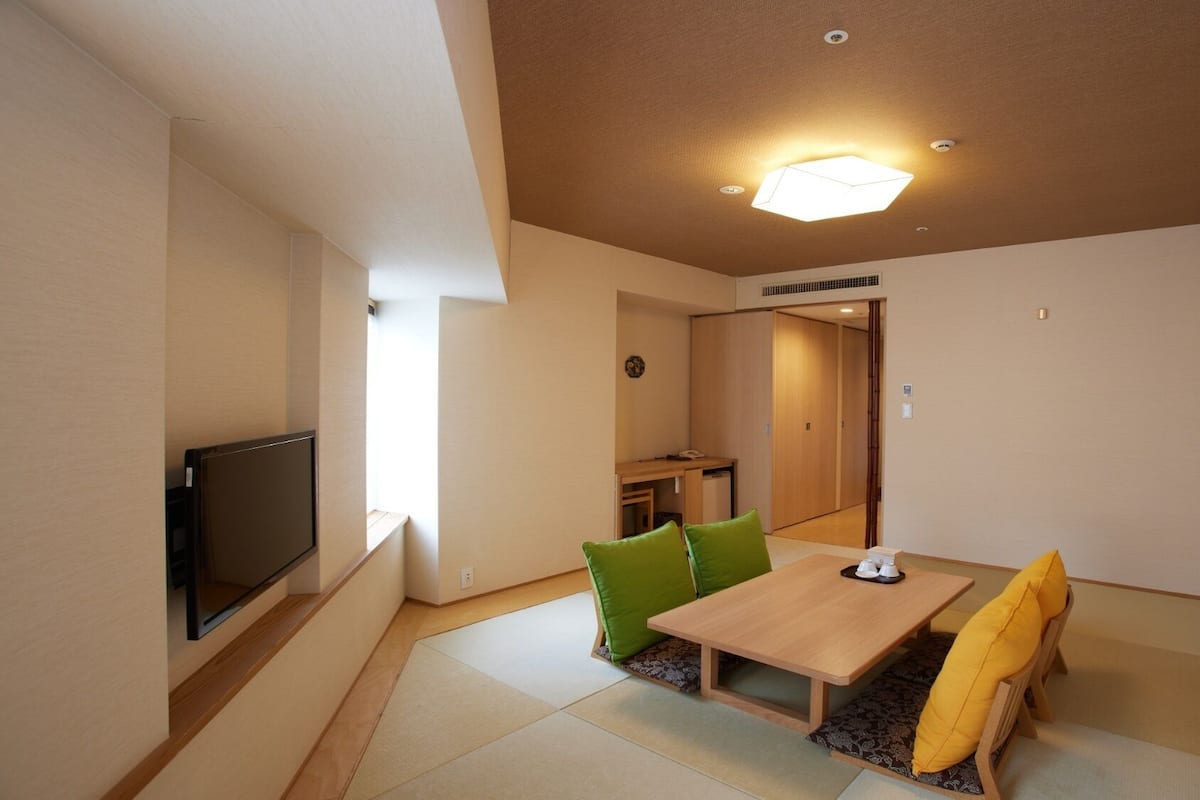 A room with a TV, a wooden table, green and yellow cushions, and a ceiling light.