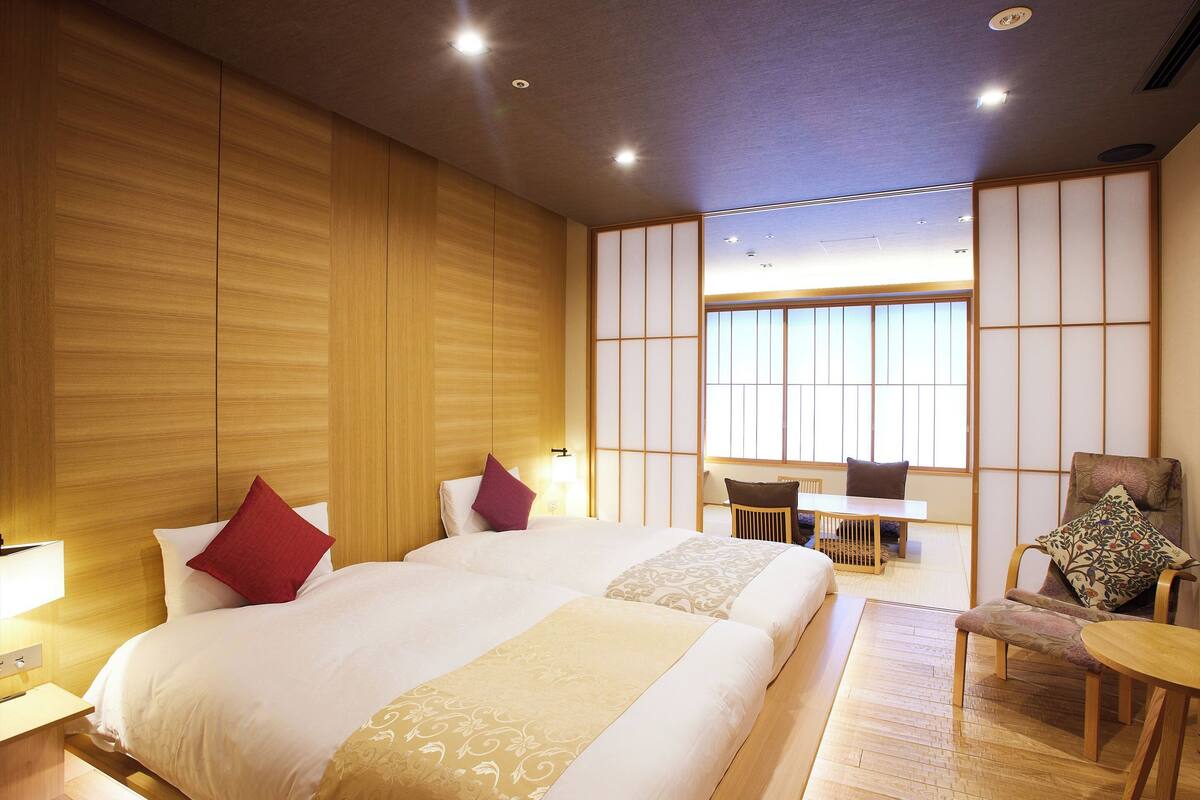 A hotel room with a large bed, a small table, a chair, and a view of the outside through a window.