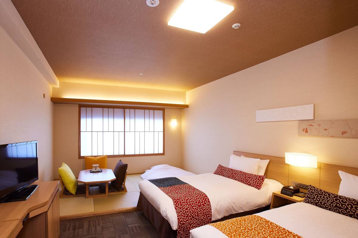 A hotel room with two beds, a desk, a TV, and a window with blinds.