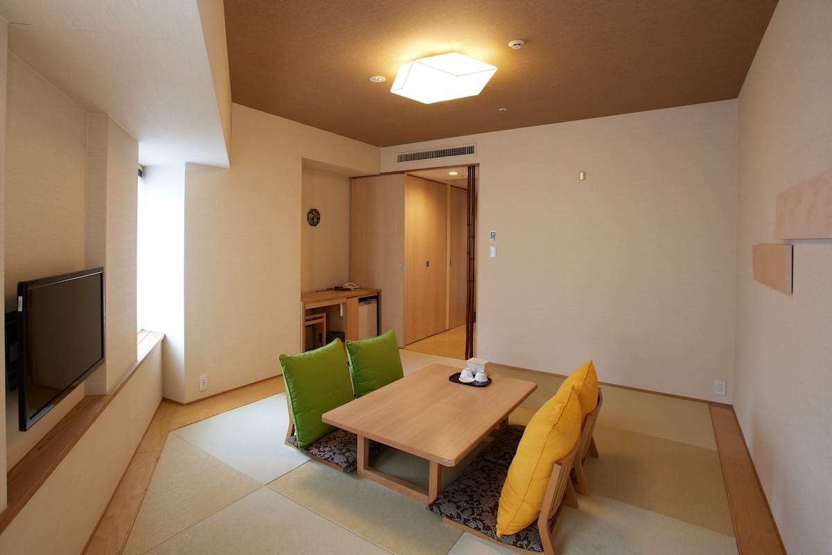 A room with tatami flooring, a low wooden table, green and yellow cushions, a television, and a ceiling light fixture.