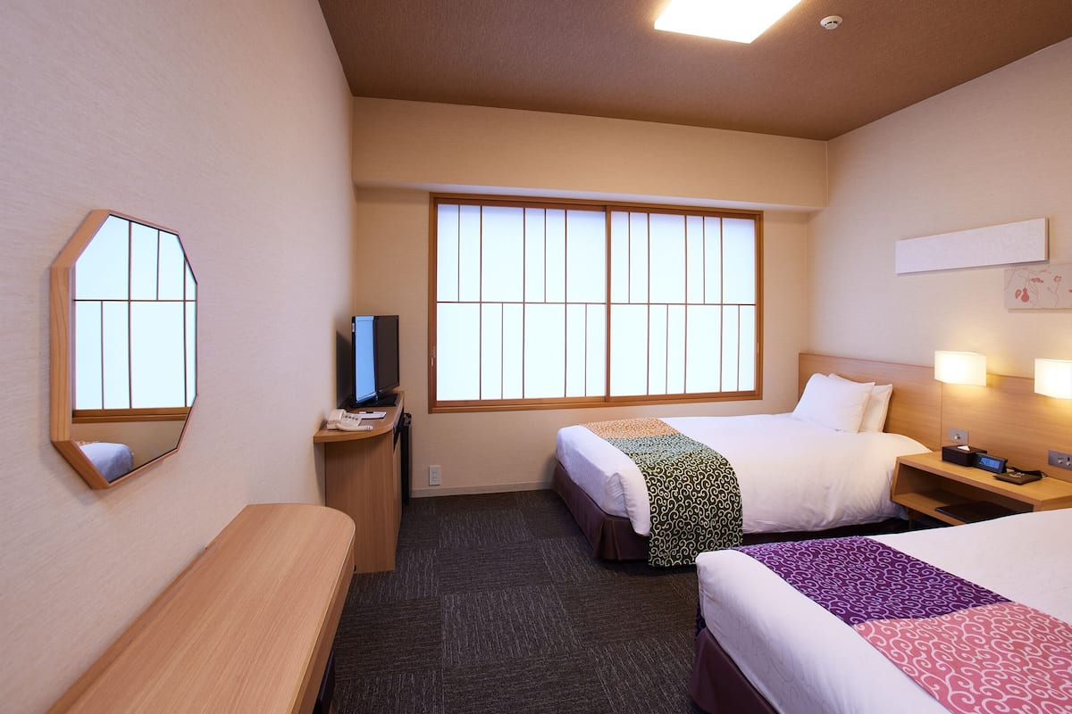 A hotel room with two beds, a desk with a computer, a large window, and a mirror.