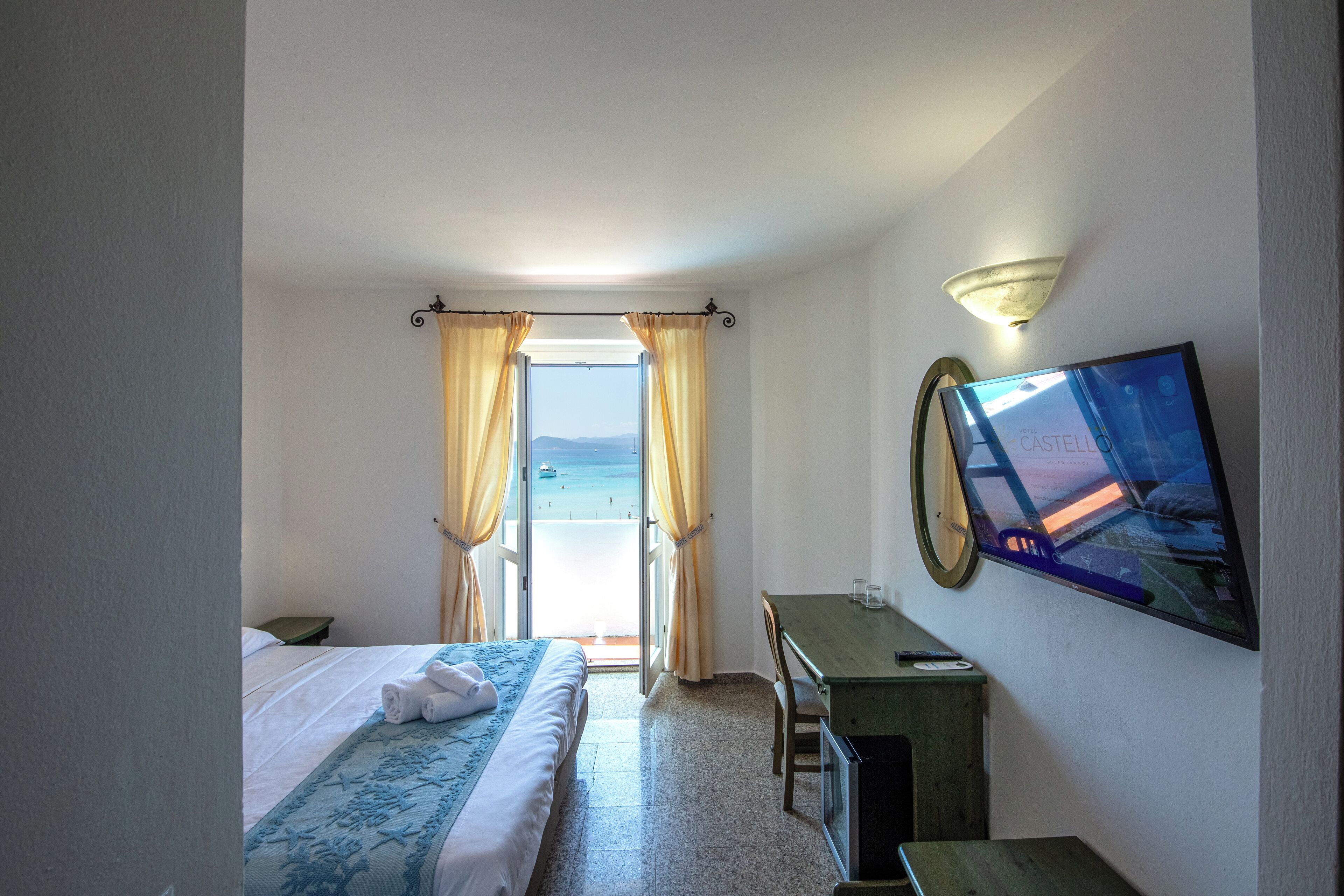 Ett hotellrum med en säng, ett skrivbord, en stol, en tv och utsikt över havet.
