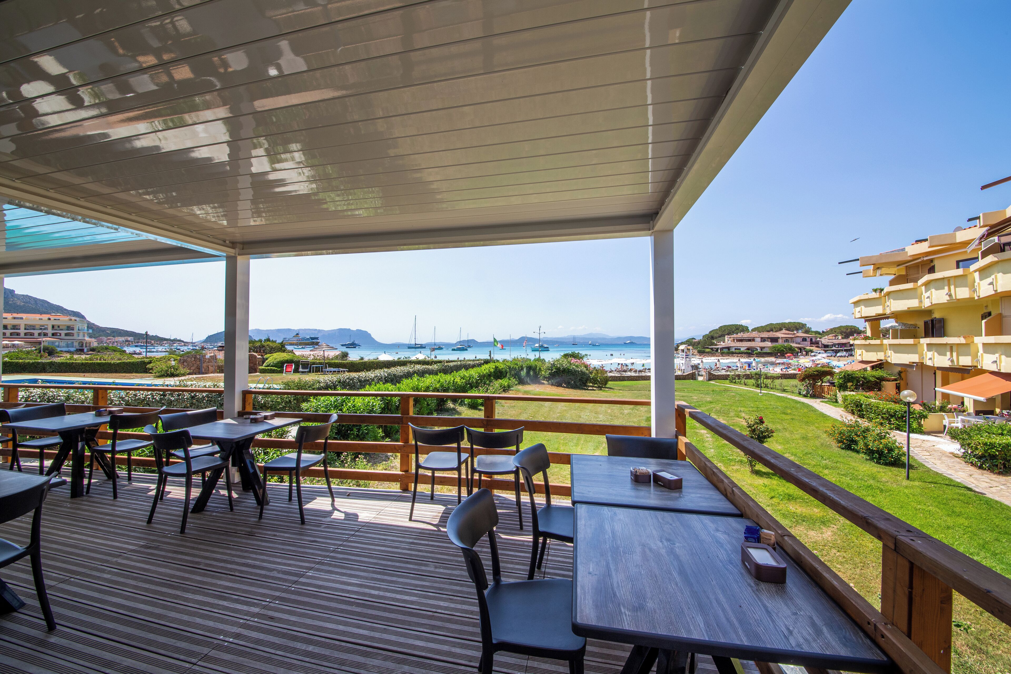A covered outdoor dining area with tables and chairs, overlooking a coastal landscape with buildings and a view of the sea.
