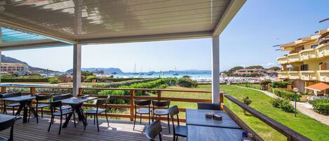 A covered outdoor dining area with tables and chairs, overlooking a coastal landscape with buildings and a view of the sea.