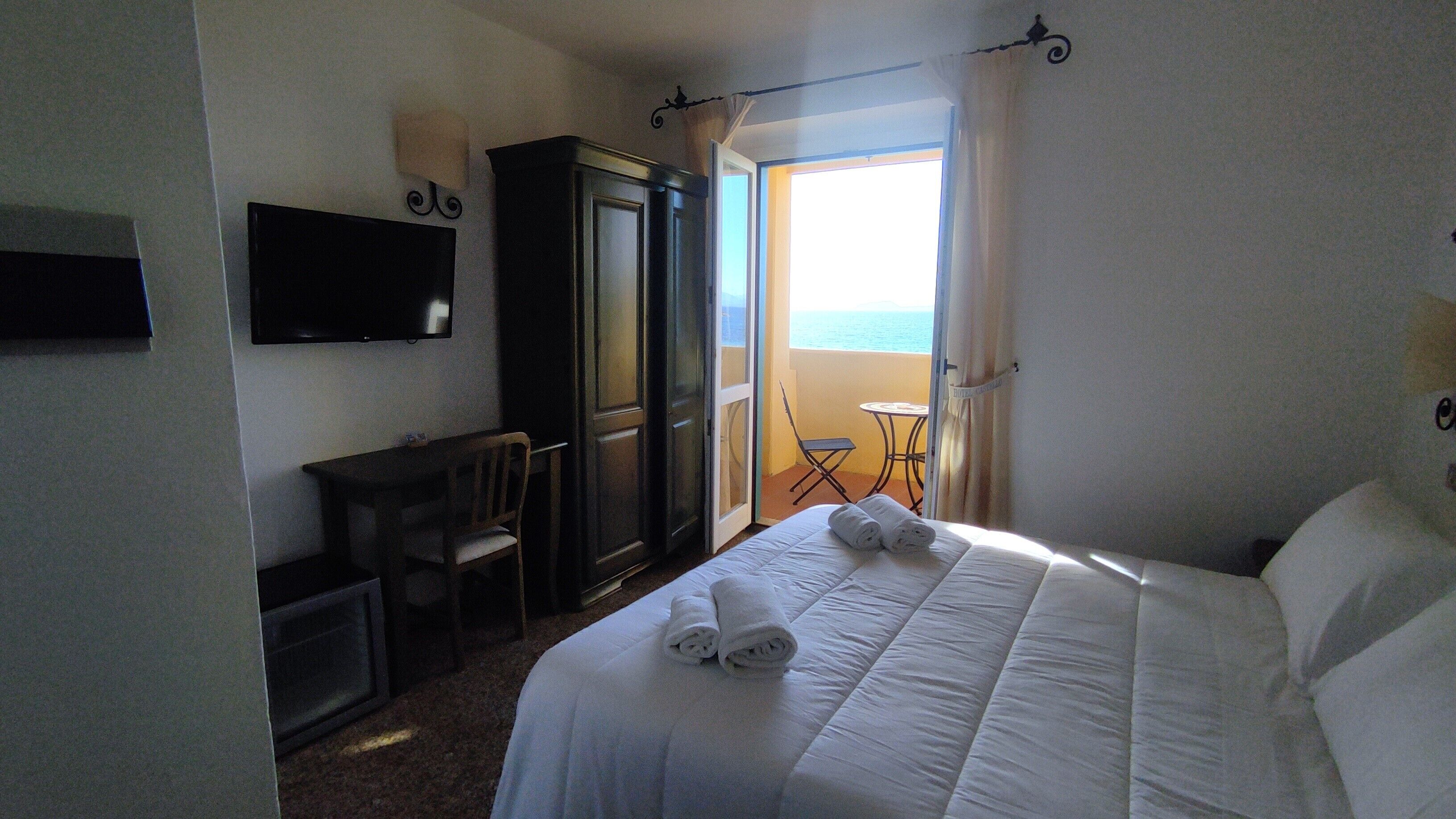 A hotel room with a bed, a desk, a chair, a television, and a balcony with a view of the sea.