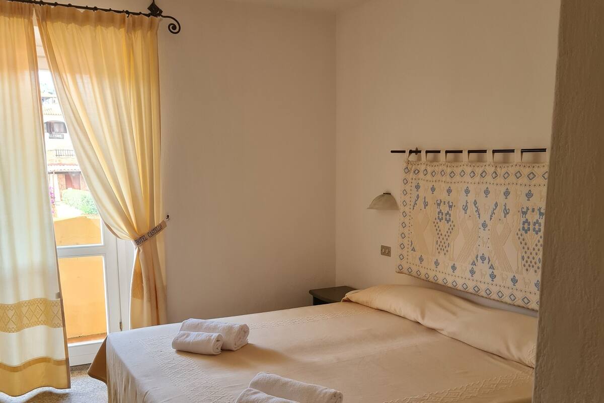 Habitación de hotel con cama, dos toallas, mesita de noche, lámpara y ventana con cortinas.
