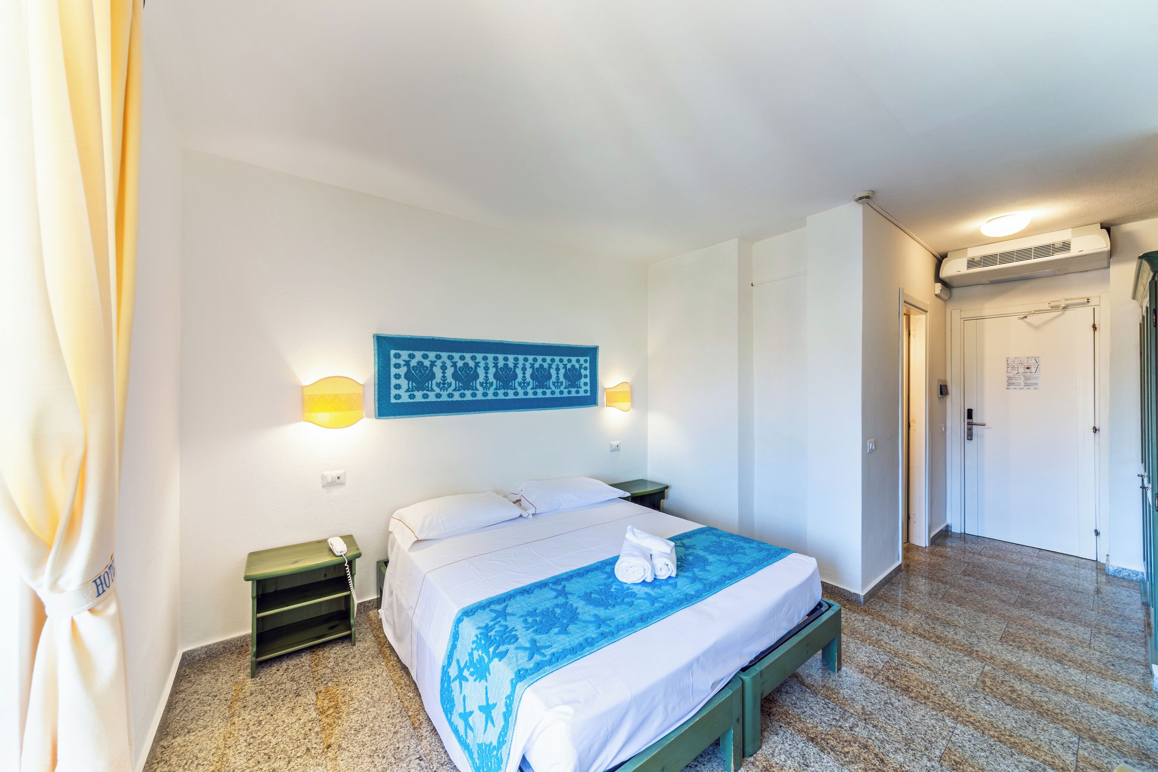 A hotel room with a bed, a nightstand, a wall-mounted lamp, a towel on the bed, and a door leading to another room.