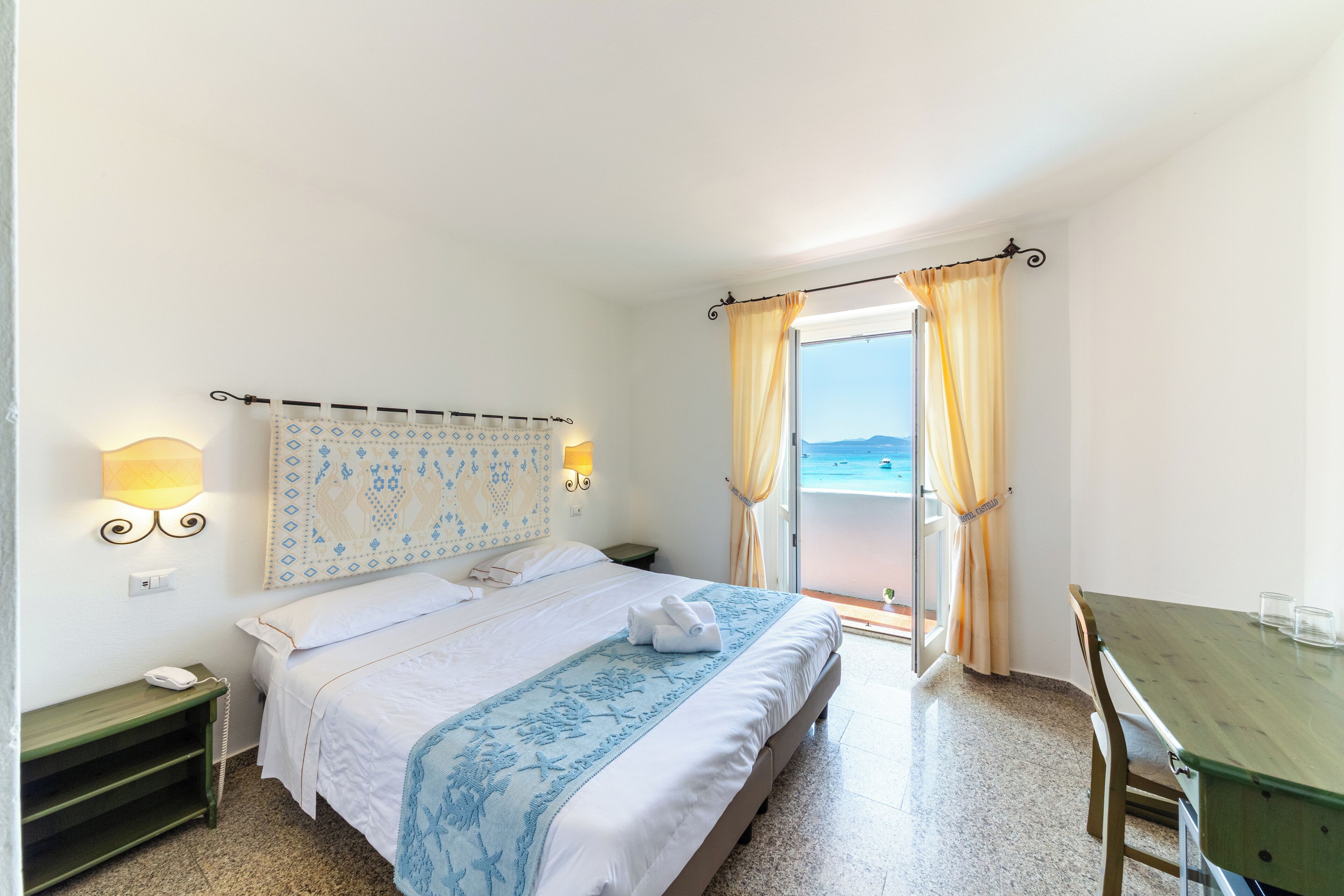 A hotel room with a bed, a bedside table, a lamp, a window with curtains, and a view of the sea.