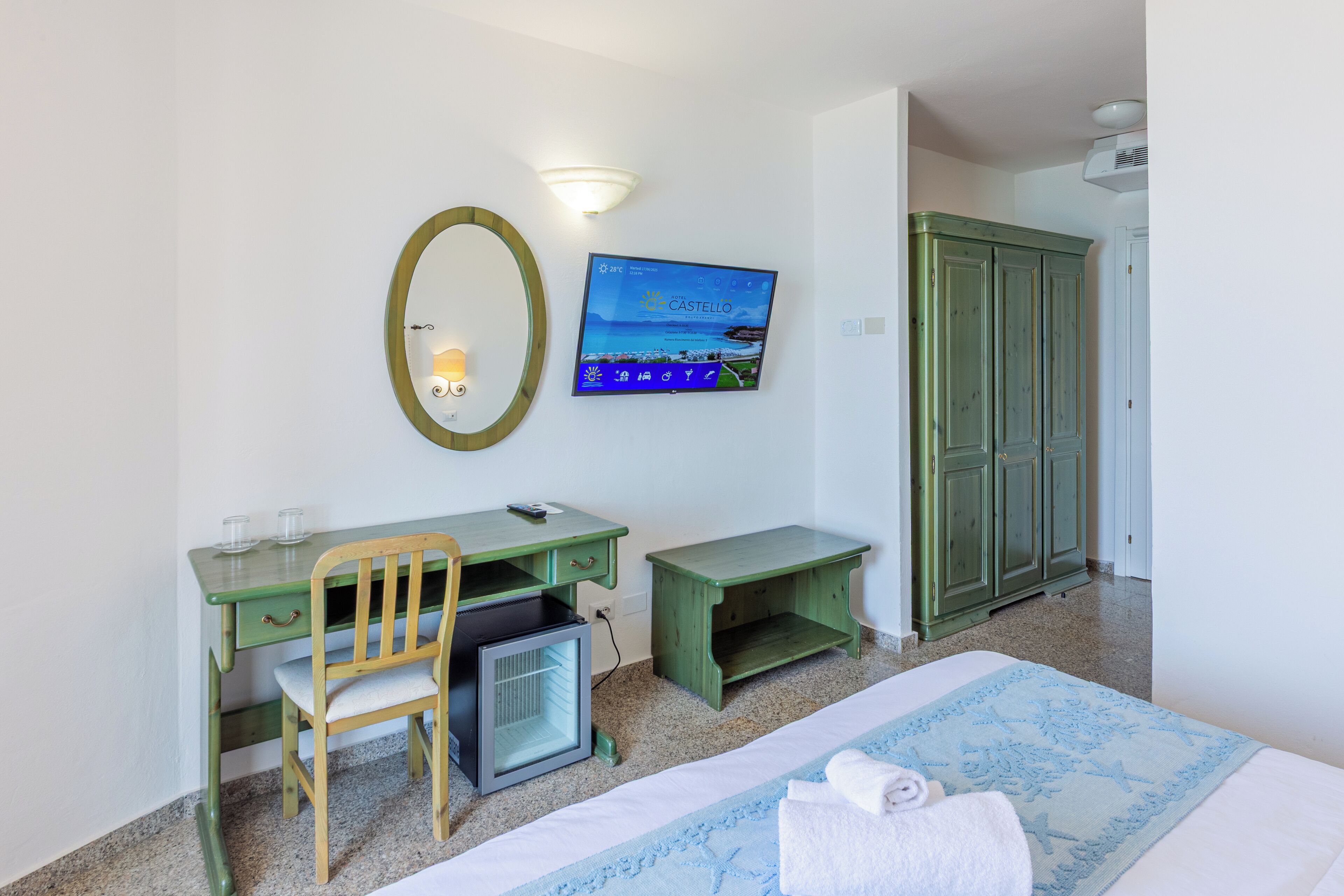 A hotel room with a bed, a desk with a chair, a small refrigerator, a television mounted on the wall, and a wardrobe.
