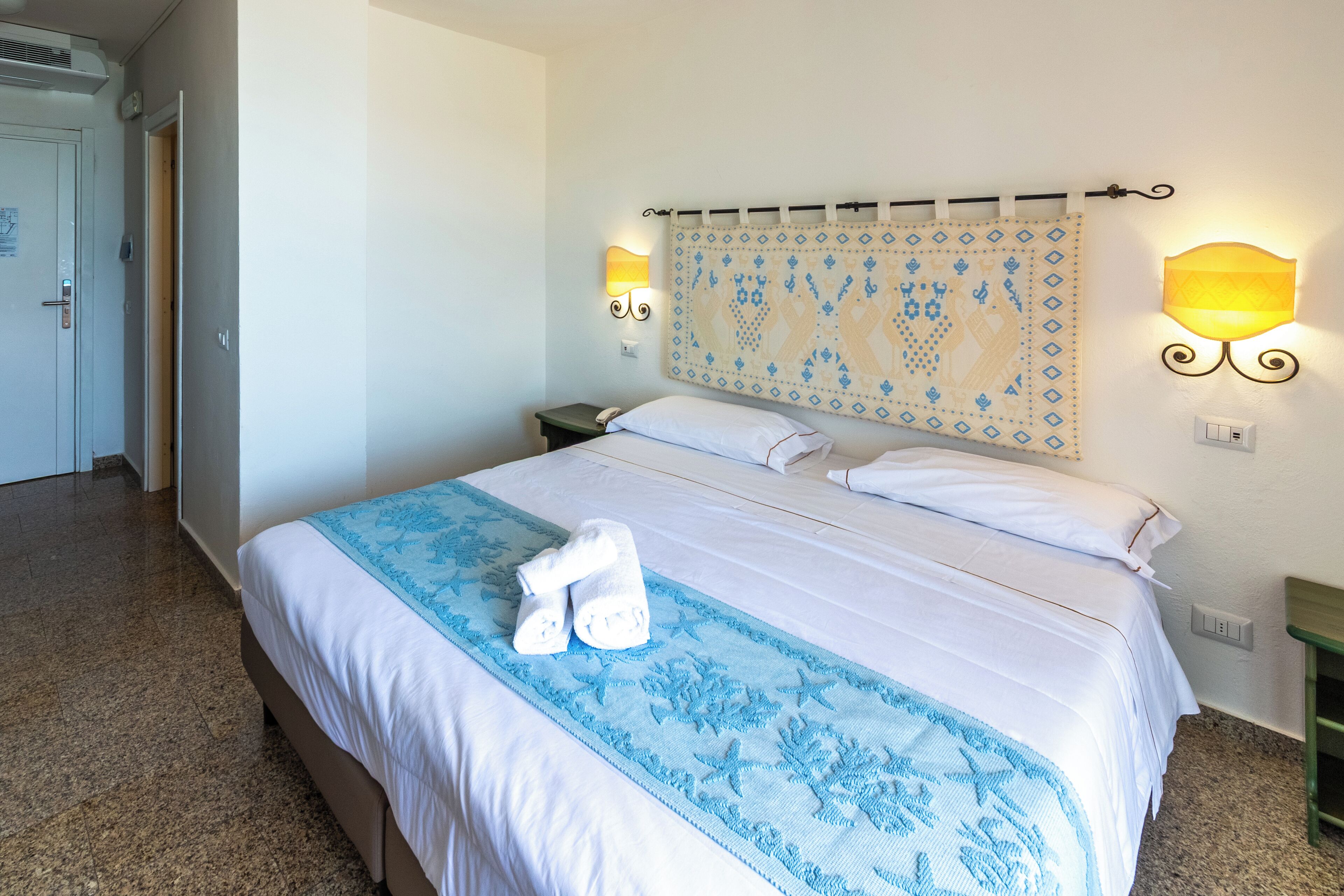Un lit bien fait, recouvert d’une couvre-lit à motifs bleus et blancs, avec deux lampes fixées au mur et une tête de lit décorative.