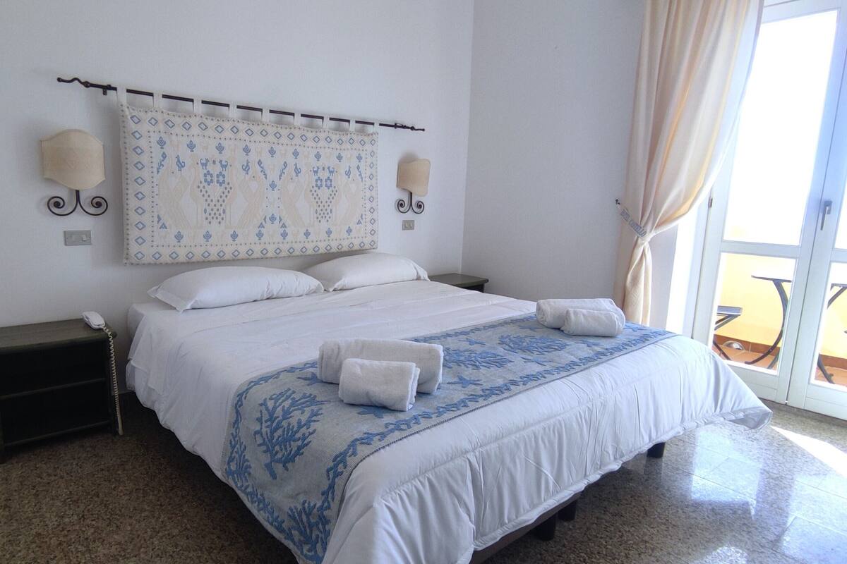 Una habitación de hotel con cama, mesita de noche, teléfono y ventana con cortinas.