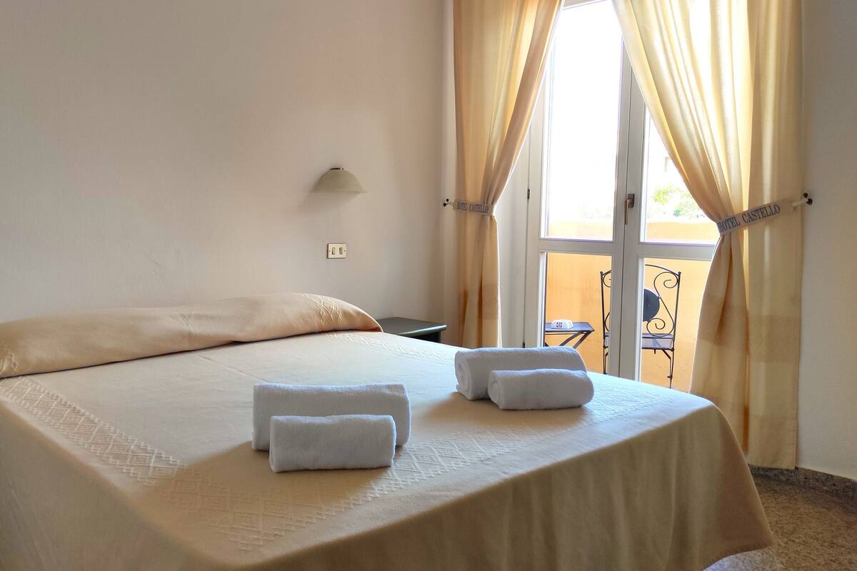 Habitación de hotel con cama, dos toallas, una mesita de noche, lámpara y vistas al exterior a través de una ventana con cortinas.