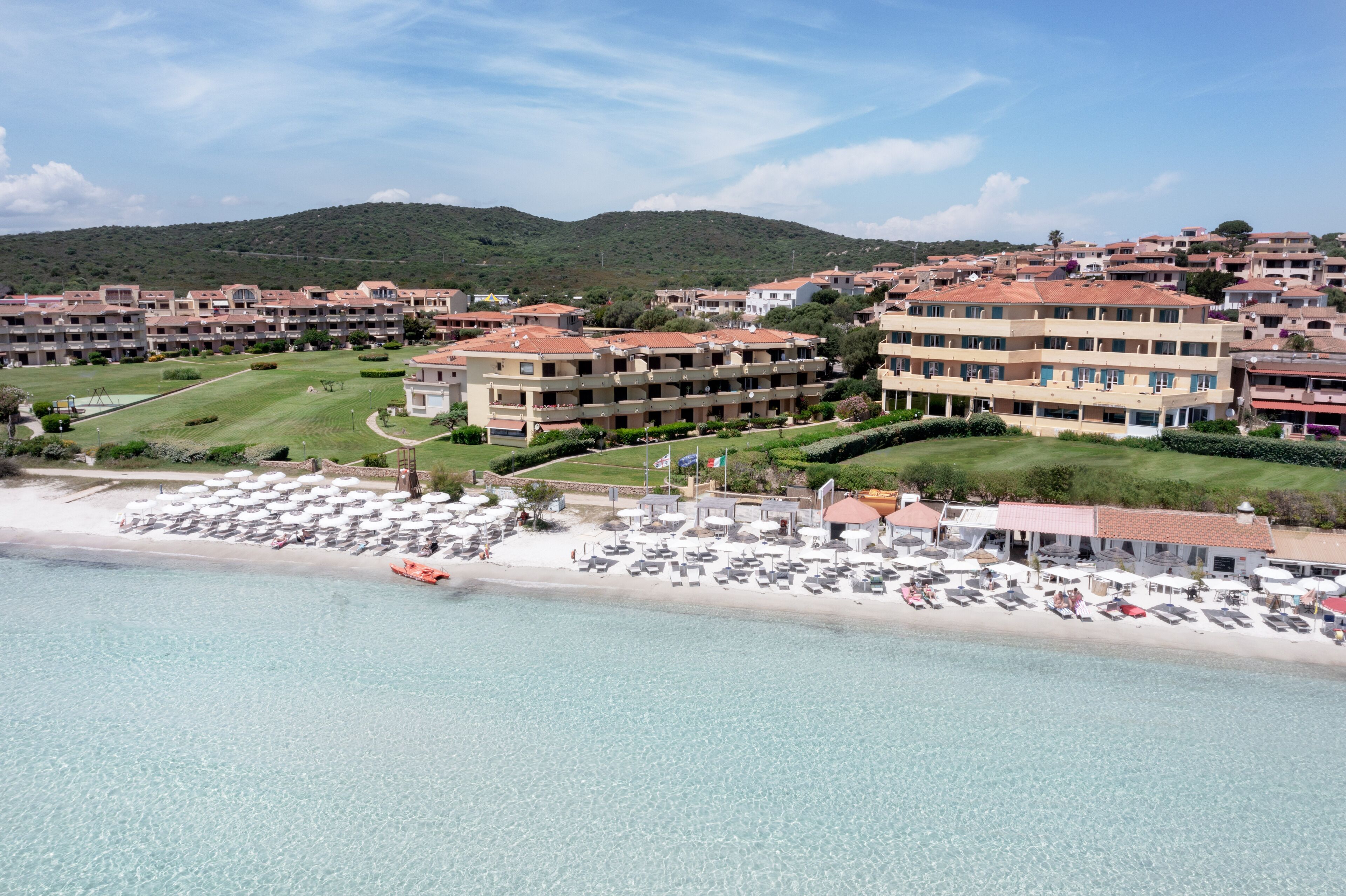 A coastal hotel with multiple buildings, a beach with sun loungers, and clear blue water.