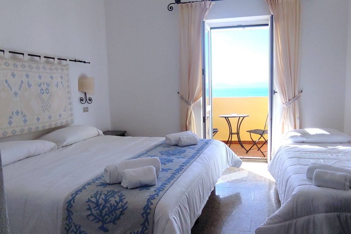 Habitación de hotel con dos camas, un balcón con mesa y sillas, y vista al mar.