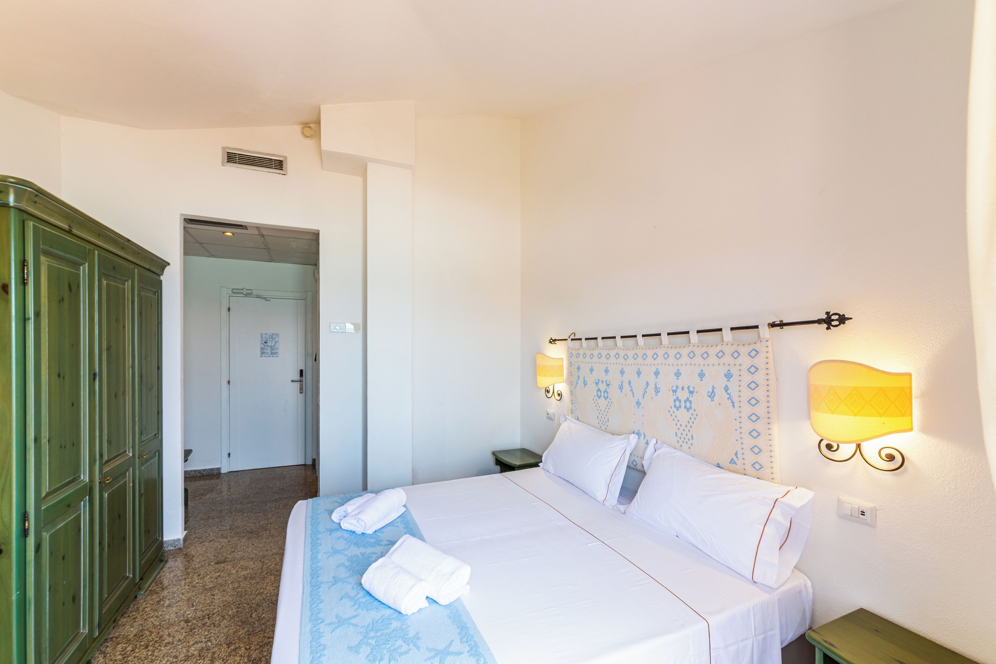 A hotel room with a bed, a nightstand, a green wardrobe, and a bathroom door.