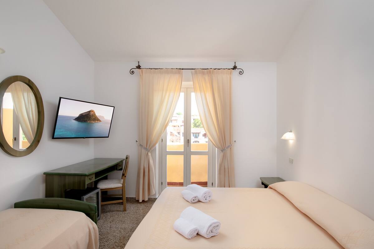 Habitación de hotel con cama, escritorio, silla, televisor y ventana con cortinas.