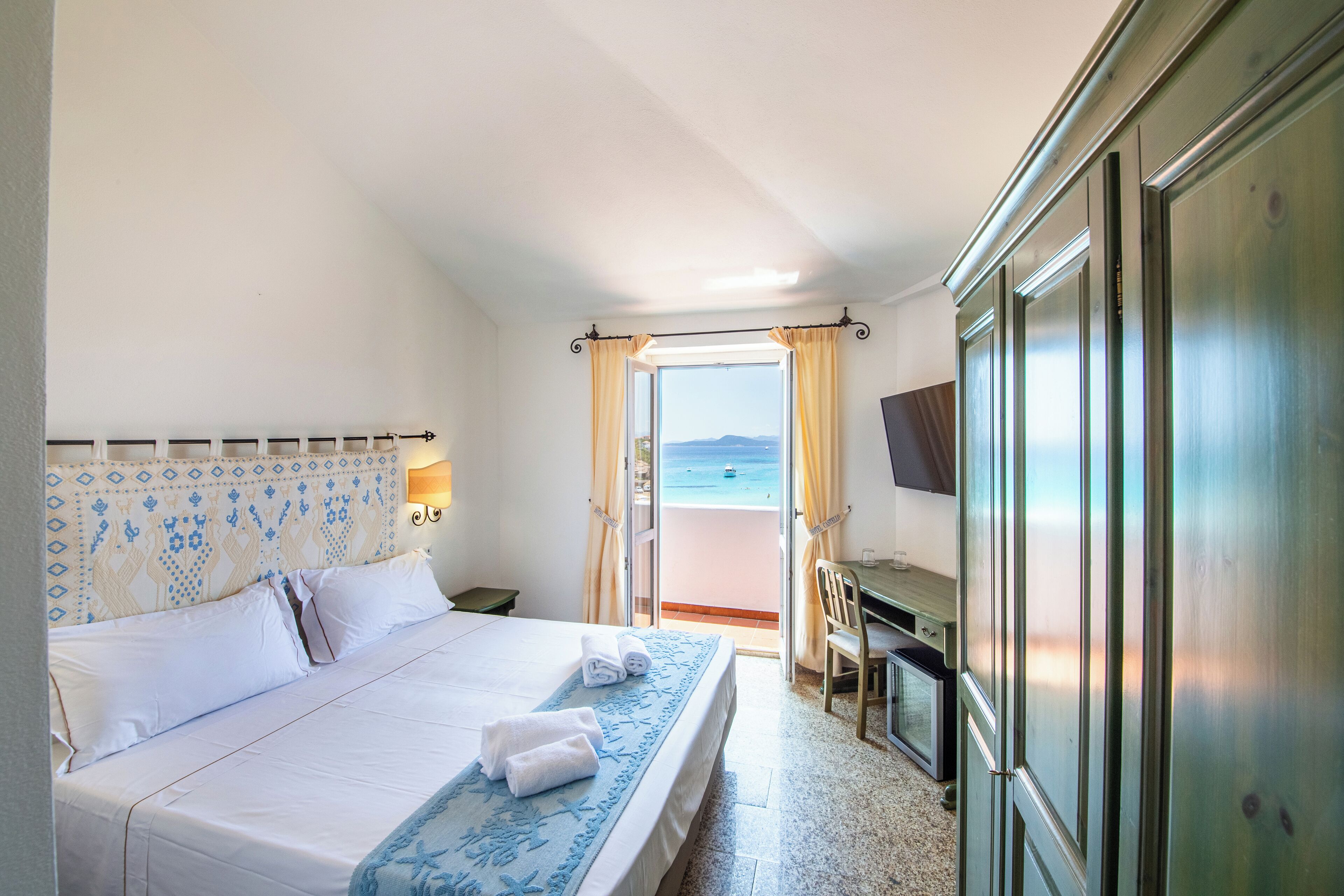 A hotel room with a bed, a desk, a chair, a TV, and a view of the sea.