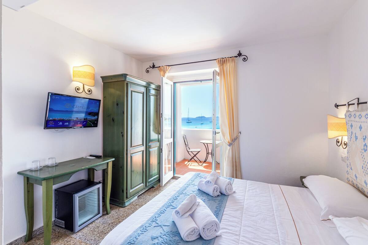 Una habitación de hotel con una cama, un escritorio con televisor, un refrigerador pequeño y vista al mar a través de una puerta abierta.