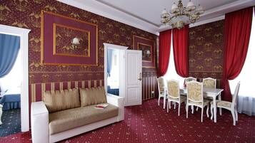 Una habitación de hotel con alfombra roja, un sofá, una mesa de comedor con sillas y un candelabro.