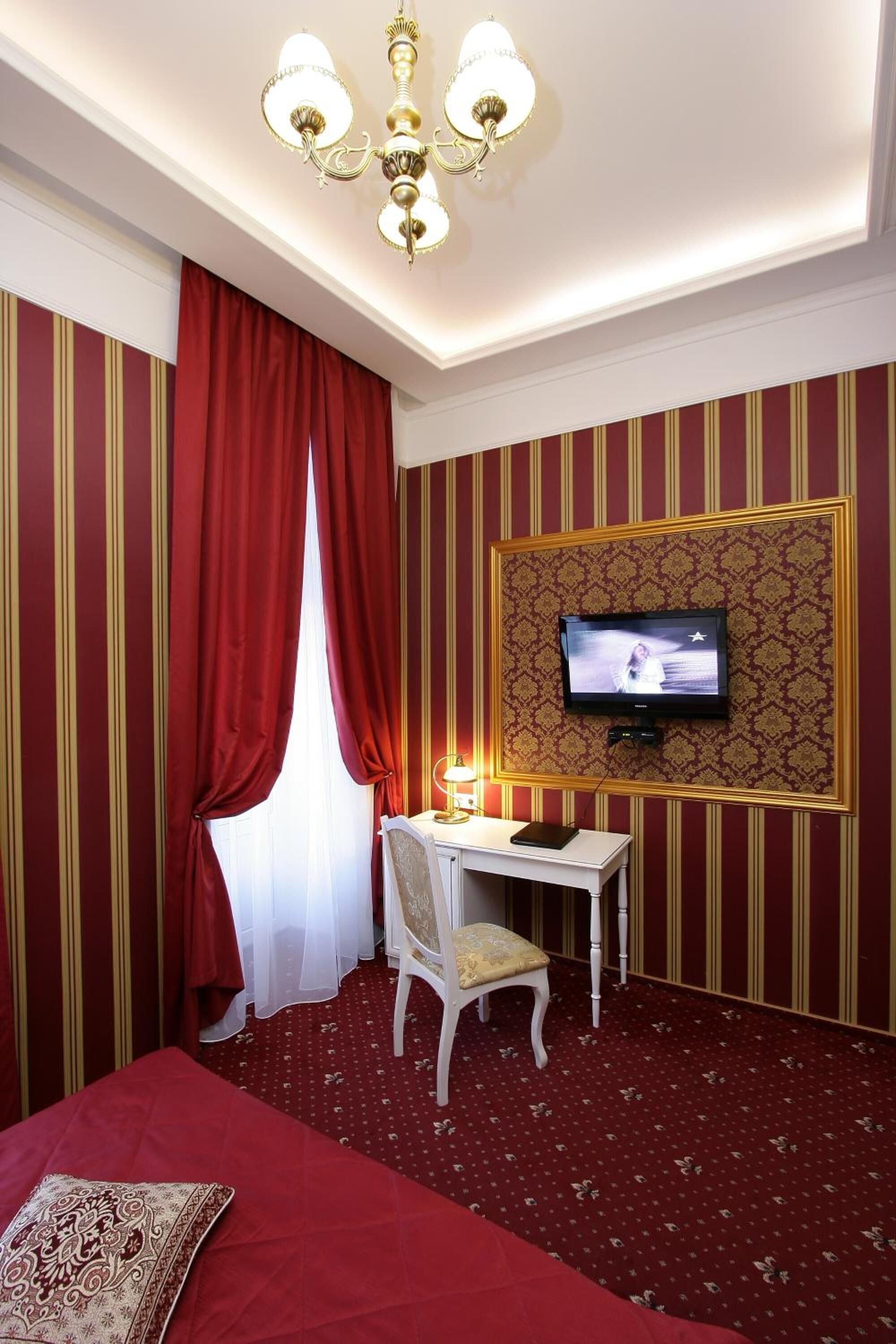 A hotel room with a bed, a desk with a chair, a television, and a chandelier.