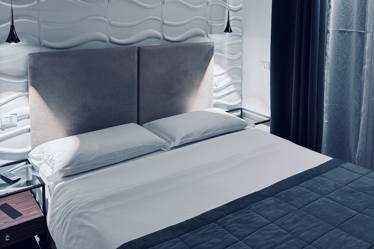 Una habitación de hotel moderna con una cama grande, un cabecero de diseño ondulado, dos lámparas colgantes y vistas al exterior a través de una ventana con cortinas.