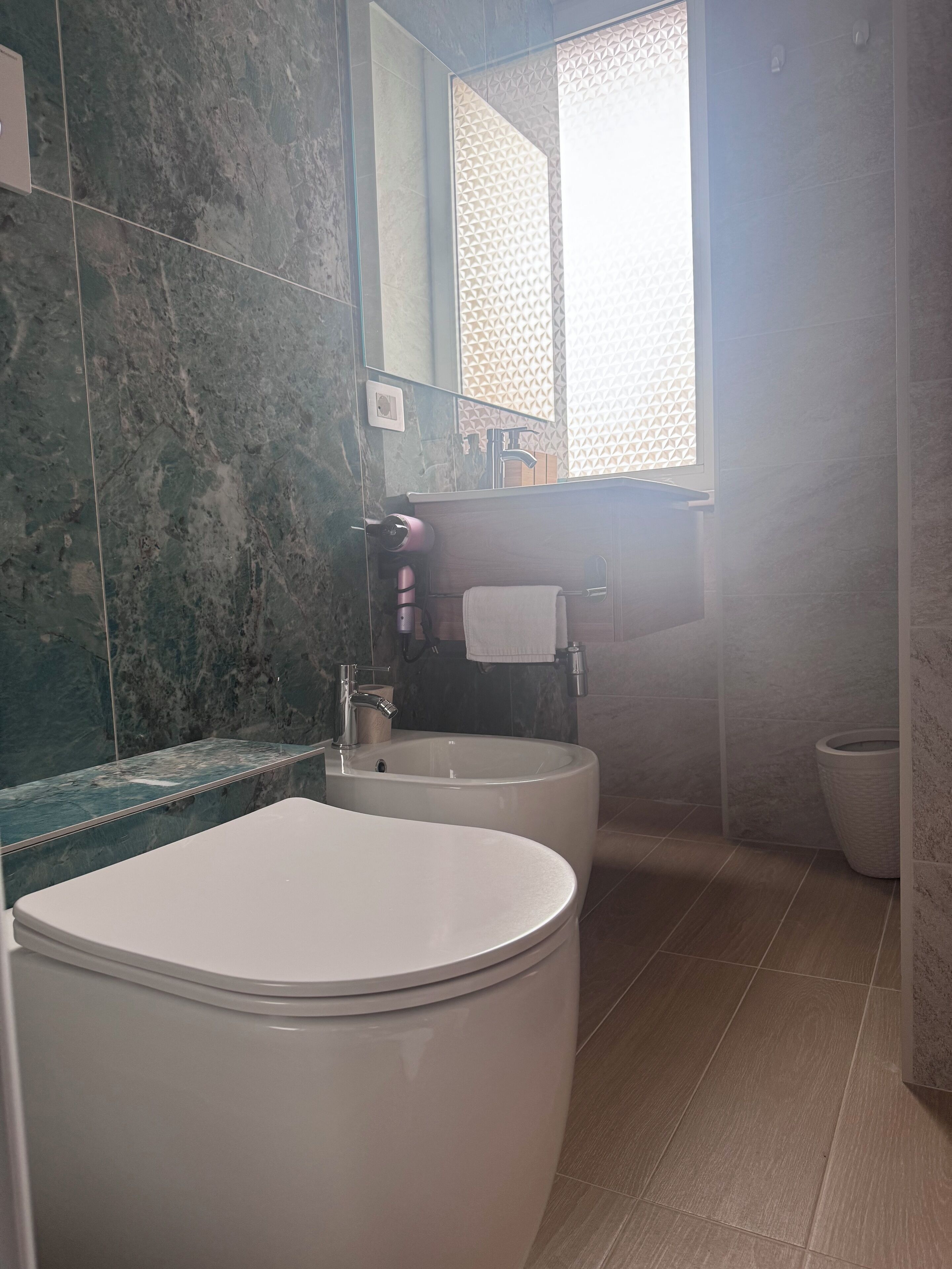 Un baño con inodoro blanco, lavamanos y bidé. Las paredes están cubiertas de azulejos con diseño tipo mármol, y hay una ventana con motivo geométrico.
