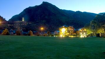 A large building with multiple floors and a prominent tower, surrounded by a grassy field and a mountain in the background.