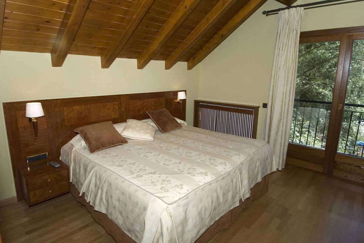 Ein Schlafzimmer mit Holzboden, einem großen Bett mit Kopfteil, Nachttischlampen und Blick auf Bäume vom Balkon aus.