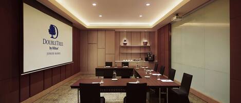 A conference room with a large screen displaying the DoubleTree by Hilton logo, a long table with chairs, and a kitchen area in the background.