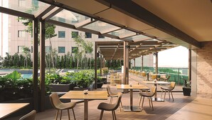 Un café al aire libre con mesas y sillas de madera, rodeado de macetas con plantas y vista a un edificio.