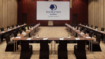 A conference room with long tables and chairs, a large screen displaying the DoubleTree by Hilton logo, and a carpeted floor.