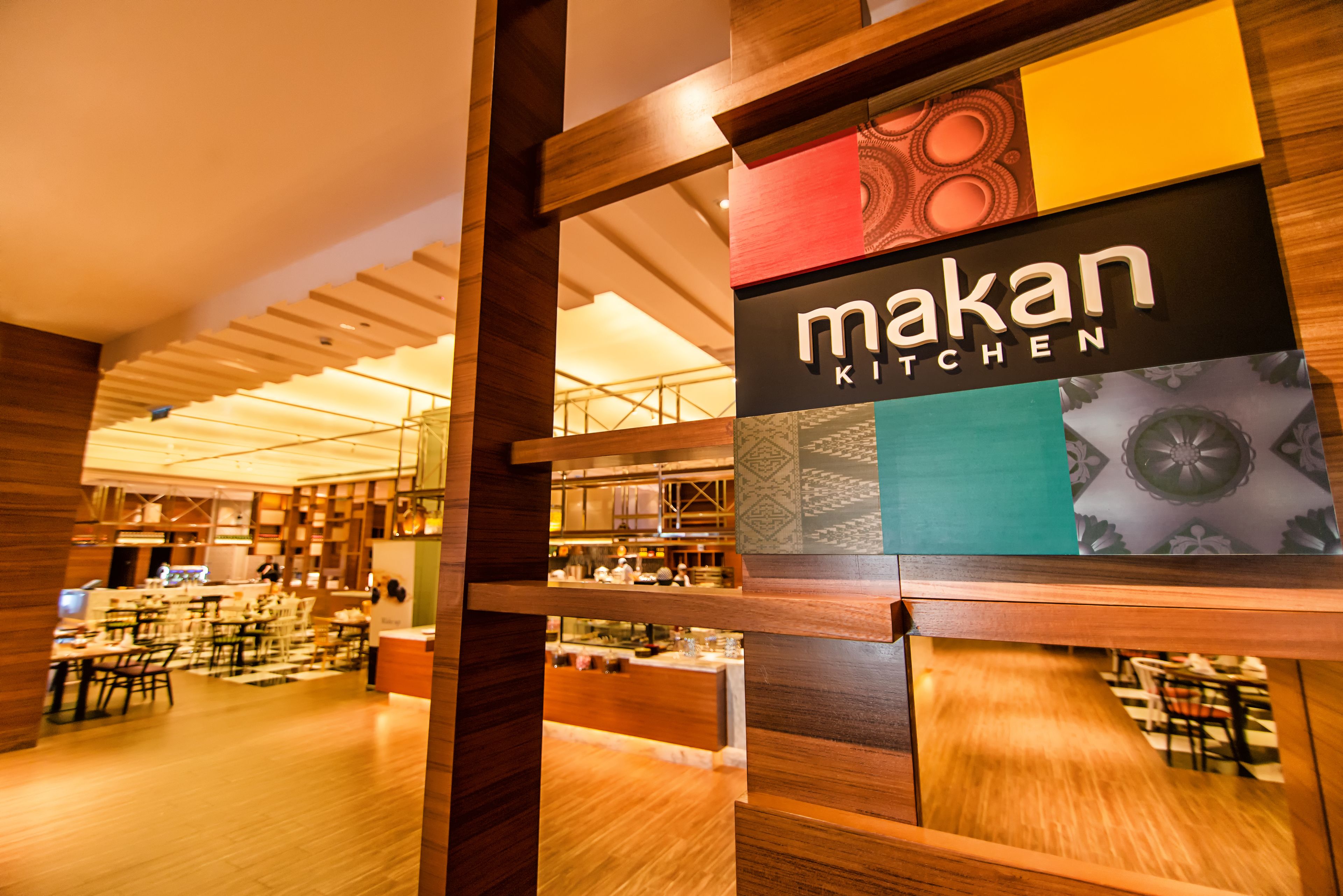 A restaurant interior with a wooden ceiling, tables, and chairs. A sign reads "Makan Kitchen".